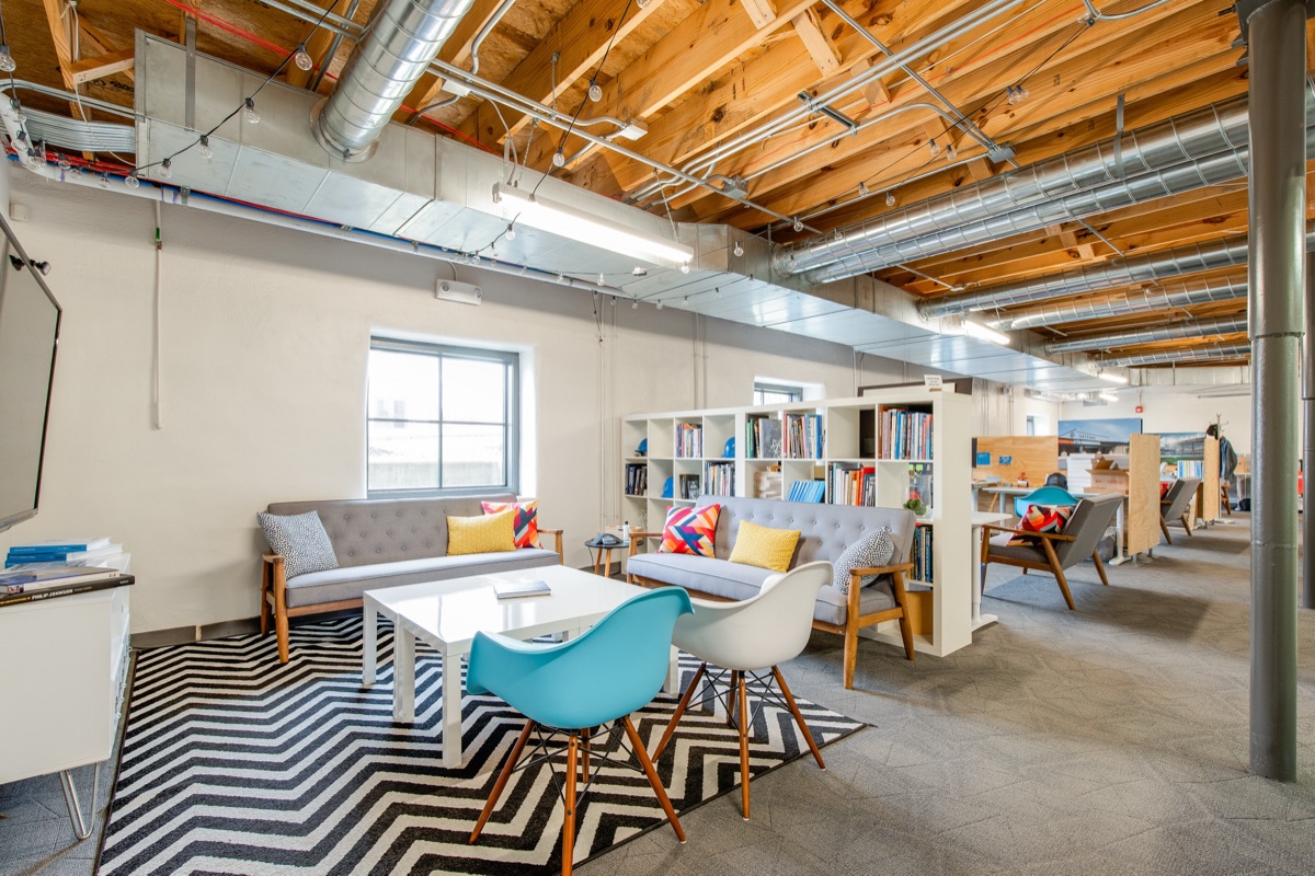 A Garden Level lounge with two grey sofas, a chevron rug and a bookshelf, under exposed ductwork and string lights.